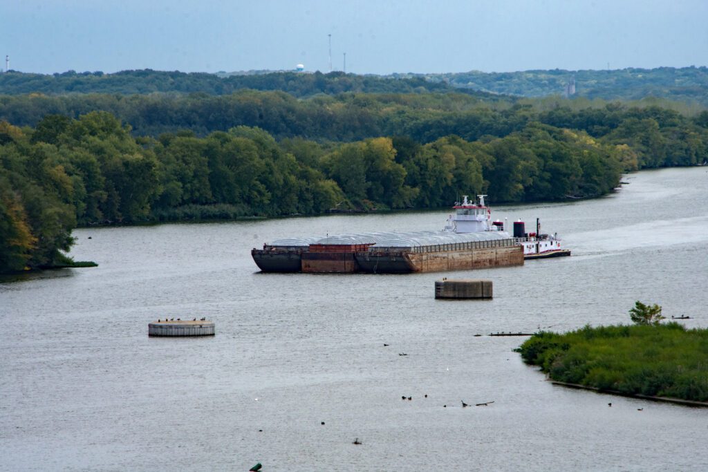 A barge travels down a wide river surrounded by dense green trees under a clear sky. Circular concrete structures are visible in the water. The serene landscape stretches into the distance. Recruiters for Barge Superintendents