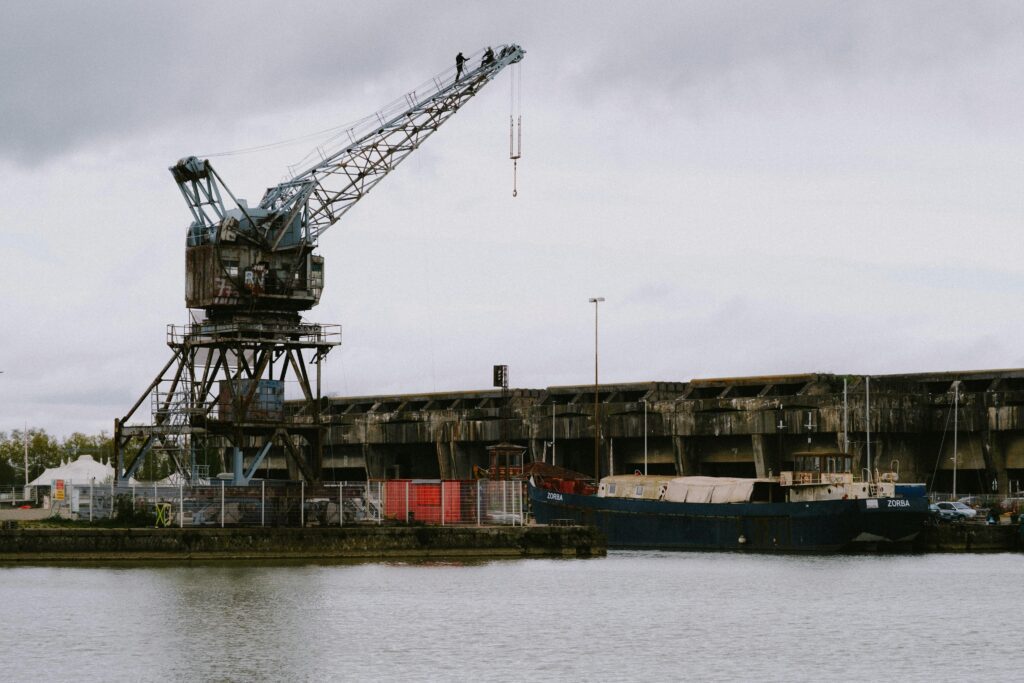 Old industrial crane by a waterfront with a docked cargo ship and large warehouse in the background. Overcast sky adds a moody atmosphere. A person stands atop the crane, adding a sense of scale. Recruiters for Port Engineers