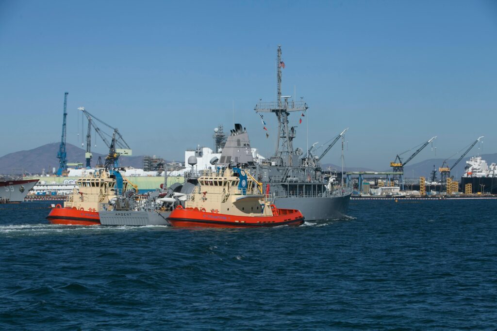 Two tugboats guide a large naval ship in a busy industrial harbor. Cranes and docked vessels are visible in the background, with a clear blue sky overhead. Recruiters for Port Operations