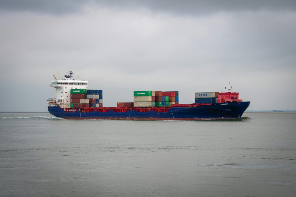 A large cargo ship with multiple colorful shipping containers is sailing on a calm sea under a cloudy sky. The ship is moving from left to right, with water ripples visible around the vessel. Recruiters for Boiler Service Engineers
