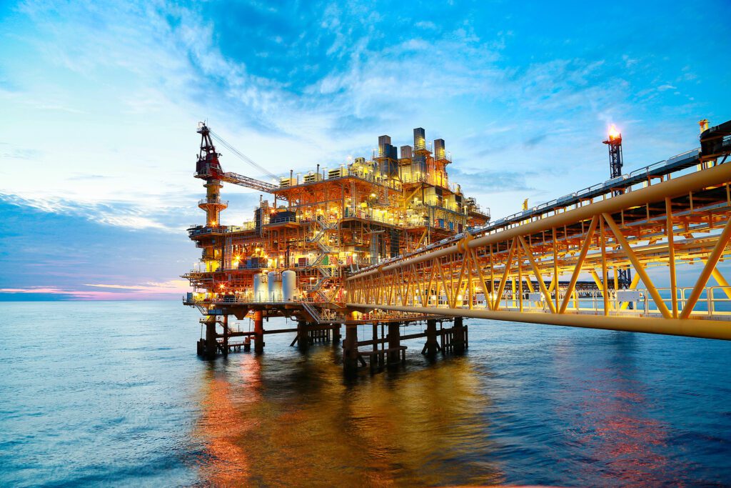 Large offshore oil rig with multiple structures and cranes, illuminated against a twilight sky. The rig is connected by a long bridge structure over calm ocean waters reflecting the warm lights from the rig, set against a vibrant blue and orange sky.; Marine Offshore Recruitment