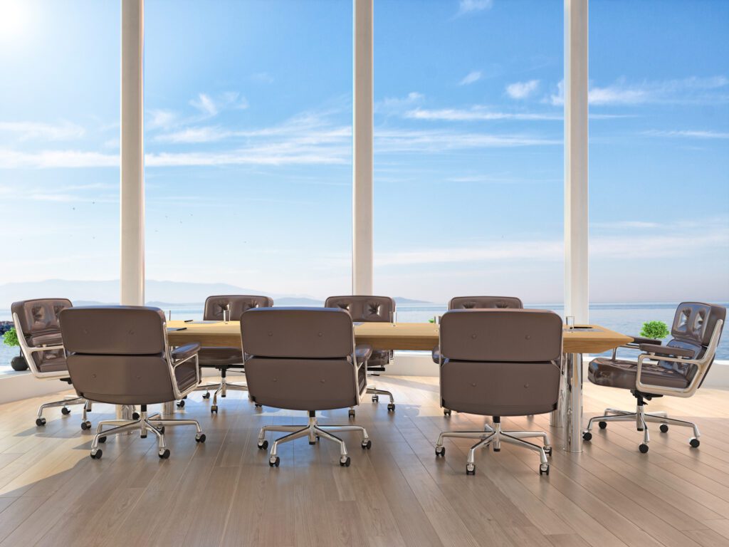 A spacious conference room with a large table and sleek brown chairs set around it. The room features floor-to-ceiling windows with a view of a clear blue sky and distant mountains, creating a bright and open atmosphere. Recruiters for Charterers