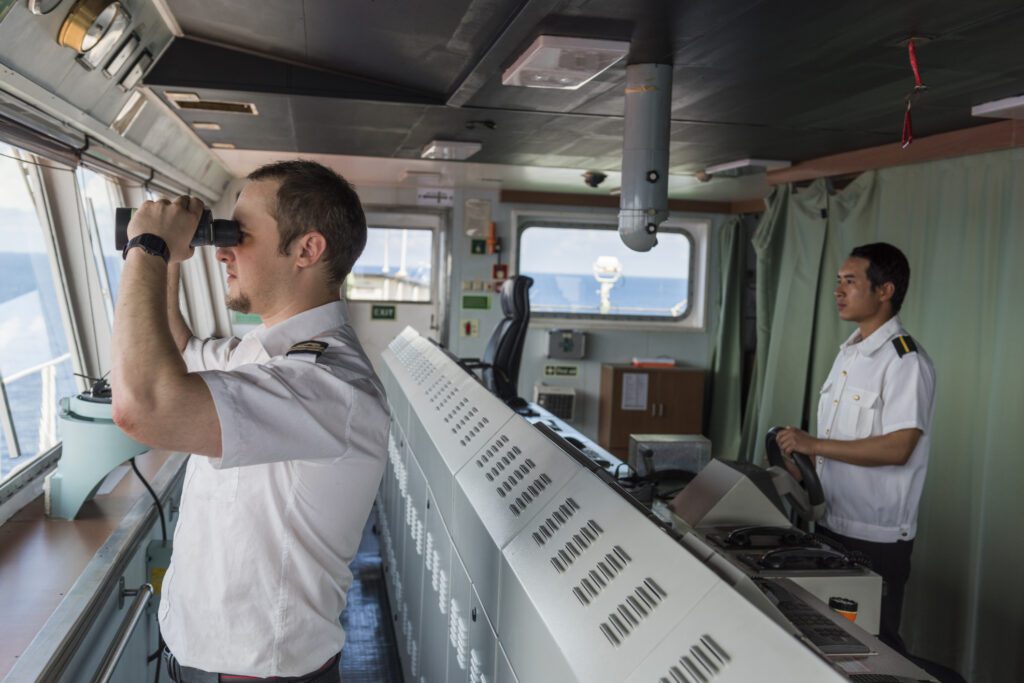 Two ship officers in uniform are on the bridge of a vessel. One is looking through binoculars out the window, while the other stands near the control panel, monitoring instruments. Ocean visible through the windows.