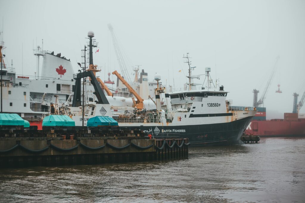 Several large ships, including a vessel labeled "Baffin Fisheries," are docked at an industrial harbor. The scene is foggy, and one ship displays a large Canadian flag. Cranes and equipment are visible in the background.
